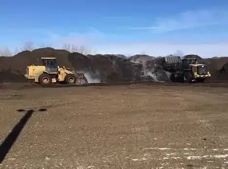 Two industrial loaders maneuver in front of large piles of soil or compost under a clear blue sky.