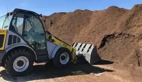 A yellow and grey front-loader machine parked next to a large pile of dark brown mulch against a clear blue sky.