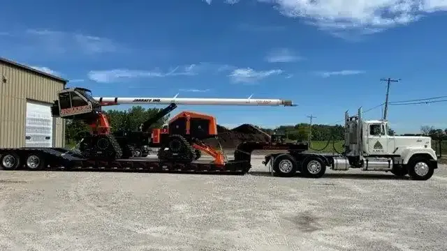 A white semi-truck hauls a flatbed trailer loaded with two pieces of orange, tracked heavy machinery on a gravel lot.