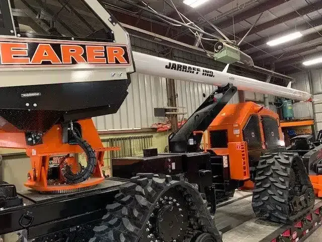 A Jarraff tree trimmer on tracks inside a warehouse, featuring an orange cab and a long white mechanical boom.