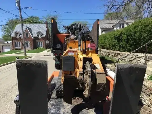 A yellow stump grinder sits on a flatbed trailer on a residential street in front of a brick house.