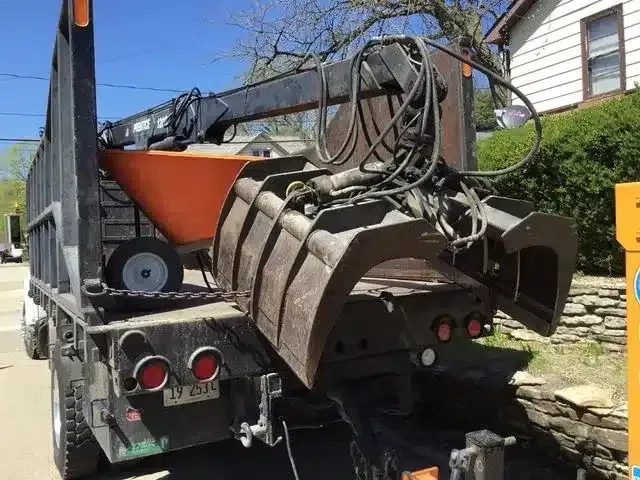 A dump truck equipped with a hydraulic grapple claw parked on a residential street near a house.