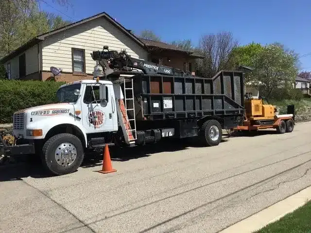 A white dump truck with a crane attachment parked on a suburban street, towing a small trailer carrying equipment.