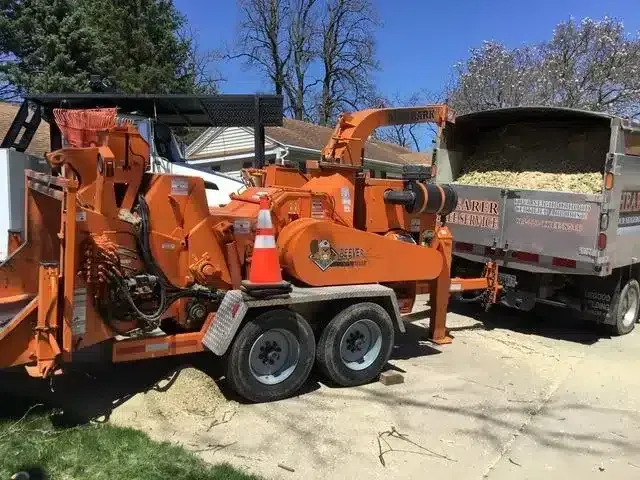 A bright orange wood chipper on a trailer, connected to a dump truck filled with wood chips, parked on a driveway.