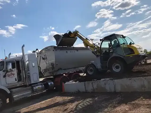 A yellow front-loader machine deposits dirt into the top of a white semi-truck trailer on a sunny, dirt construction site.