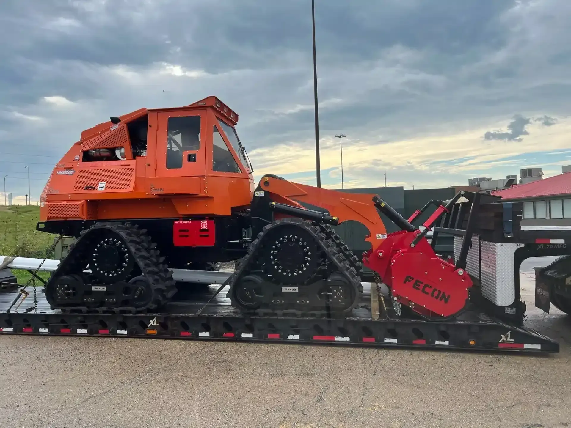 An orange Fecon tracked mulcher machine secured on a flatbed trailer outdoors under a cloudy sky.