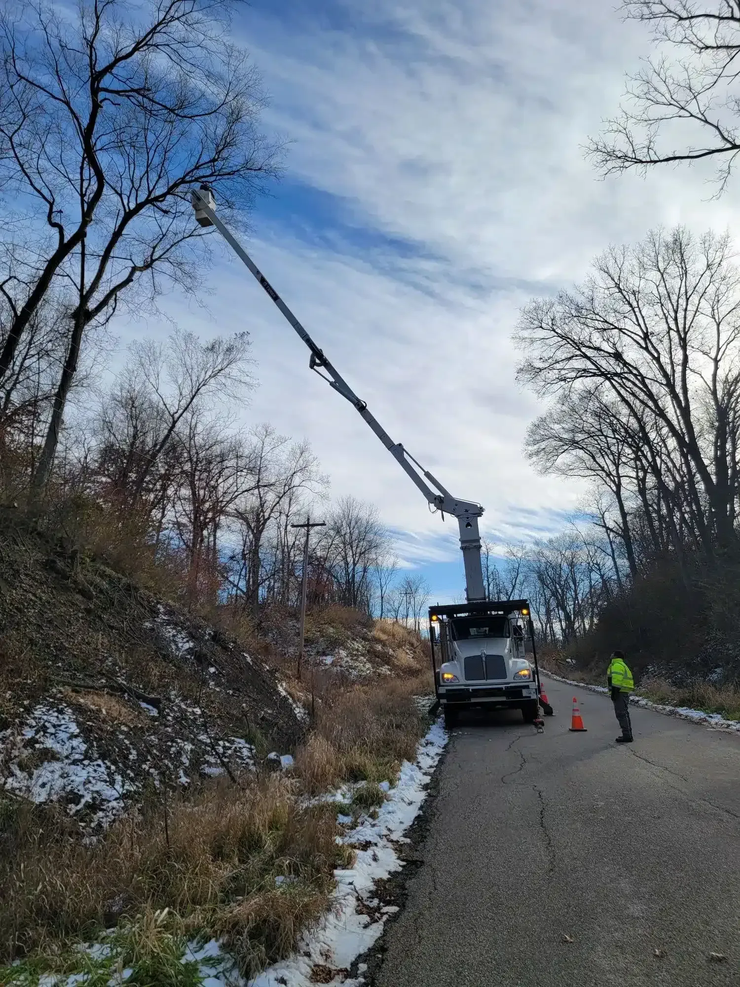 A utility truck with an extended boom lift works on trees along a paved road with a worker standing nearby.