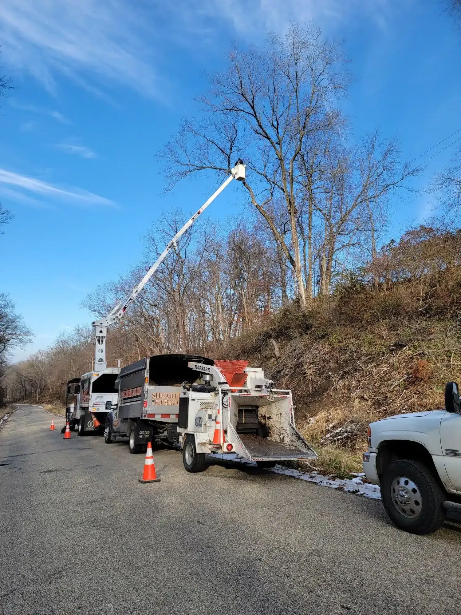 A bucket truck with an extended boom and a wood chipper parked on a gravel road, with trees against a clear blue sky.