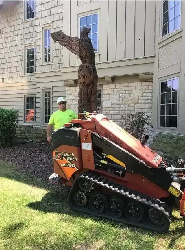 A worker in a neon green shirt stands next to a tracked stump grinder in front of a house with a large eagle wood carving.