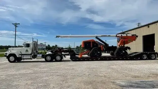 A white semi-truck hauls a trailer carrying a bright orange forestry machine on a gravel lot under a blue sky.