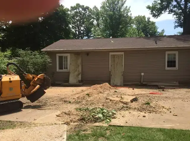 A stump grinder sits on a dirt lot in front of a tan house with two doors and windows on a sunny day.