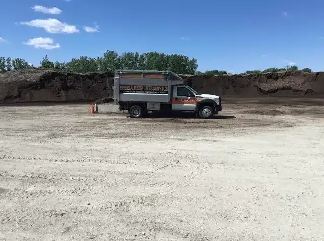 A service truck parked in an open, dirt-covered lot with large mounds of dark soil in the background under a blue sky.