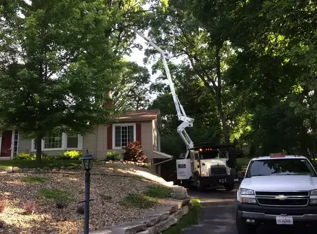 A utility truck with a raised boom arm parked in a driveway next to a house and a white pickup truck.