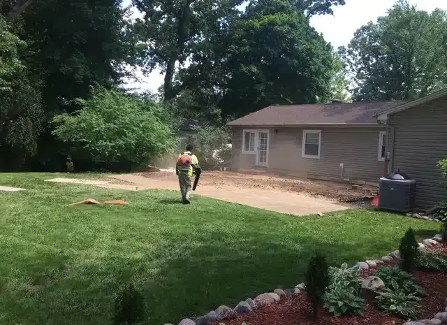 A worker in a high-visibility vest uses a leaf blower to clear debris from a dirt-covered patio area near a house.