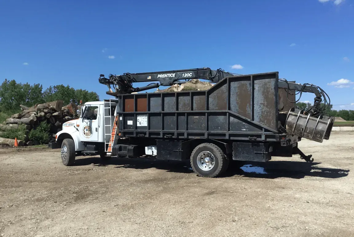 A white grapple truck with a black debris container and crane arm, parked in a dirt lot with a pile of logs nearby.