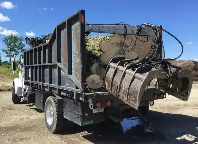 A white dump truck loaded with large logs, featuring an attached hydraulic grapple arm, parked on a dirt lot.
