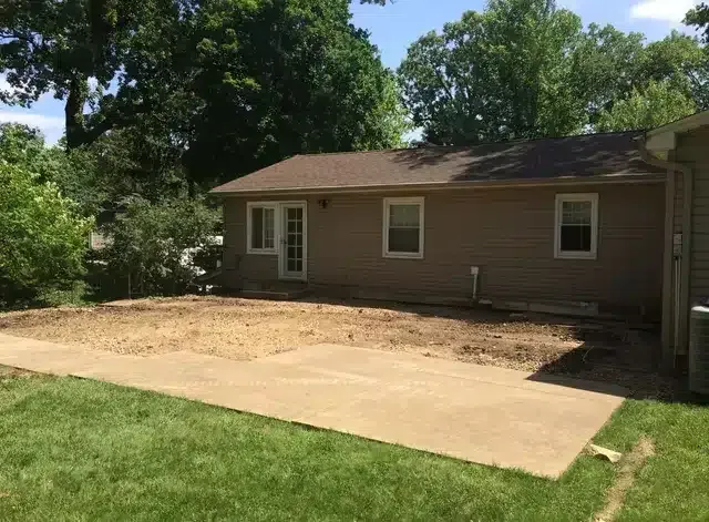 A beige house with a concrete patio in front of a dirt yard, surrounded by green trees and grass on a sunny day.