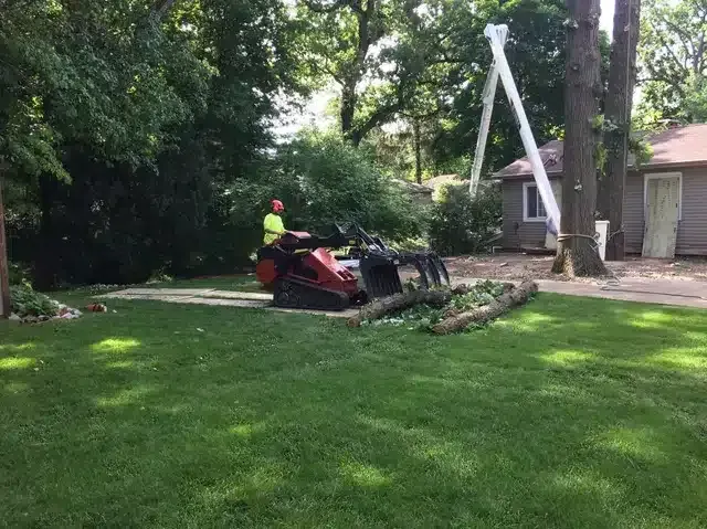 A person in a neon shirt operates a red tracked skid steer with a claw attachment to move logs in a grassy yard.