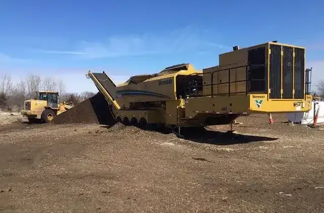 A yellow industrial Vermeer compost turner operates on a dirt field with a loader nearby under a clear blue sky.