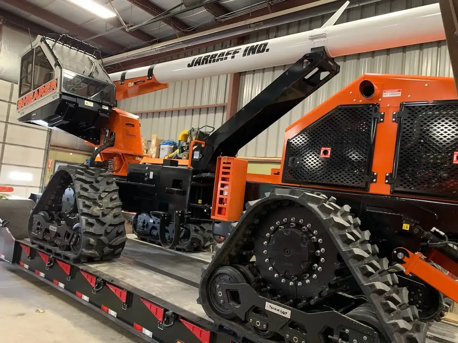 An orange and black forestry machine with large tracks sits on a flatbed trailer inside a garage.