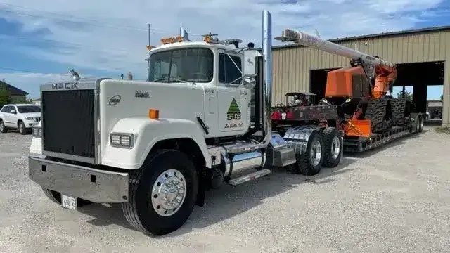 A white Mack semi-truck towing a lowboy trailer carrying an orange piece of heavy construction equipment.