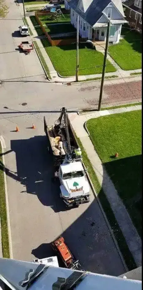 A white tree-trimming truck is parked on a residential street next to a house, with its boom extended upward.