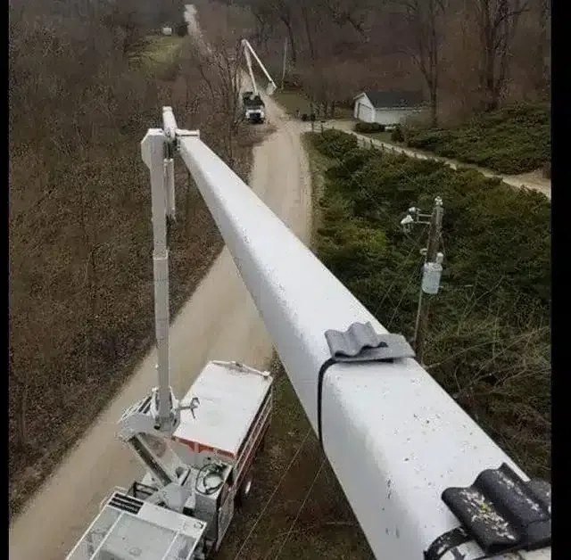 A high-angle view of a utility truck's extended boom reaching over a rural dirt road, with another truck parked nearby.