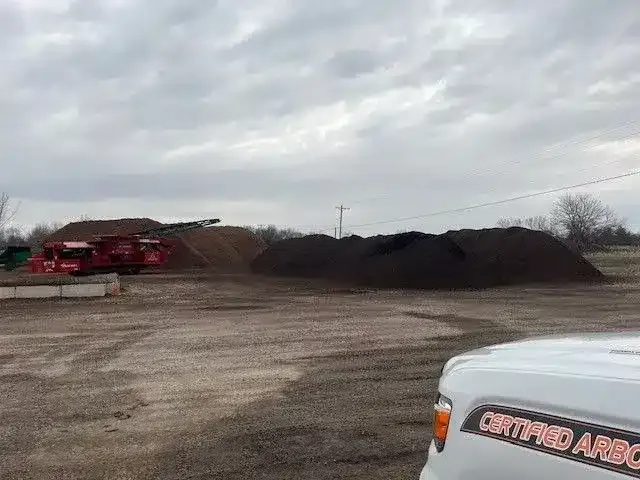 Large piles of mulch and wood chips sit in an outdoor storage yard with red processing equipment under a cloudy sky.