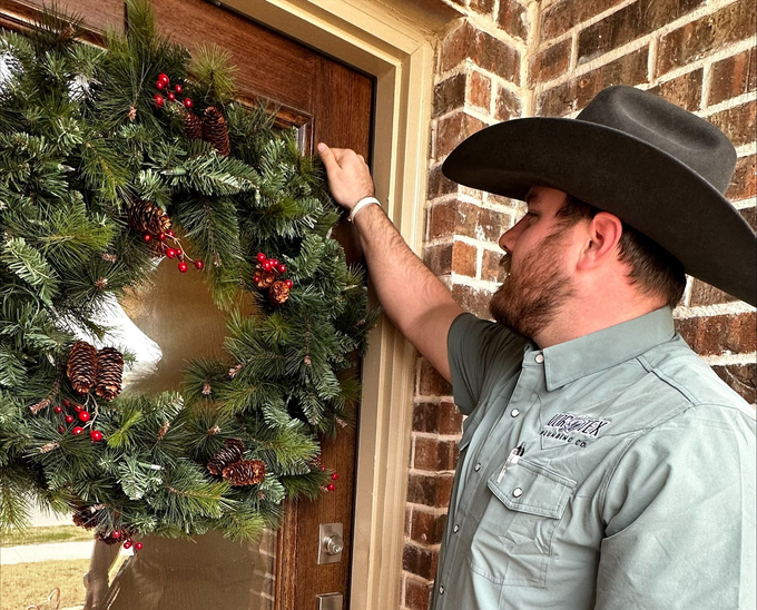 Man in cowboy hat hangs a Christmas wreath on a wooden door; brick wall.