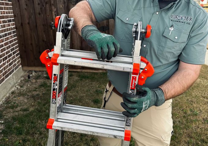 Person adjusting a foldable orange and silver ladder outdoors, wearing green gloves and a green shirt.