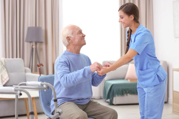 Caregiver in blue scrubs helping a seated man stand in a bright living room