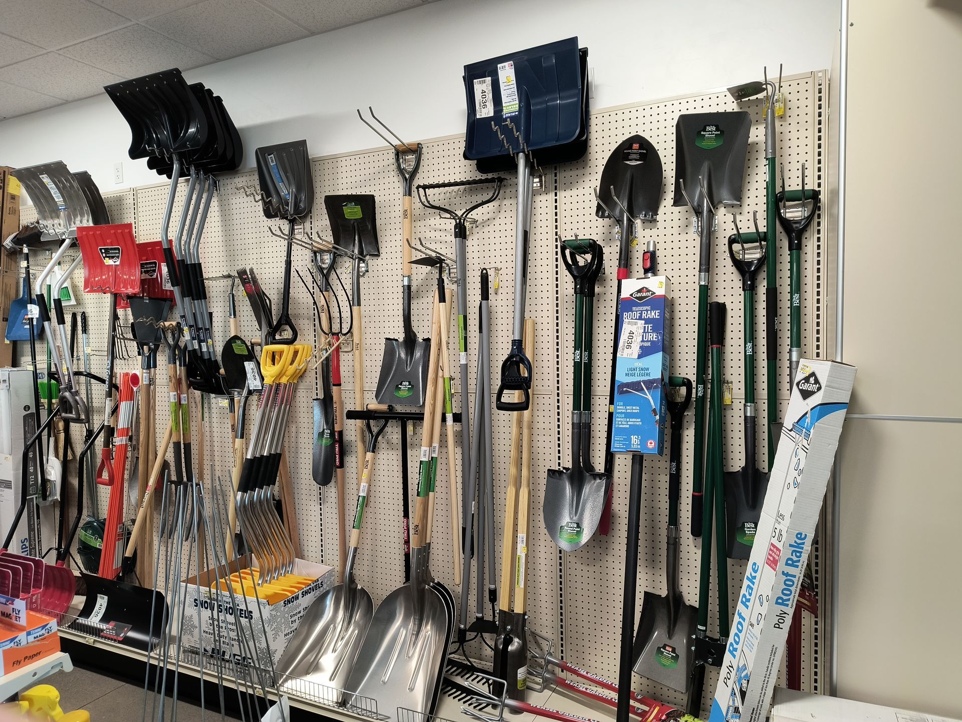 Garden tools displayed on a pegboard wall in a store. Shovels, rakes, and snow shovels are visible.