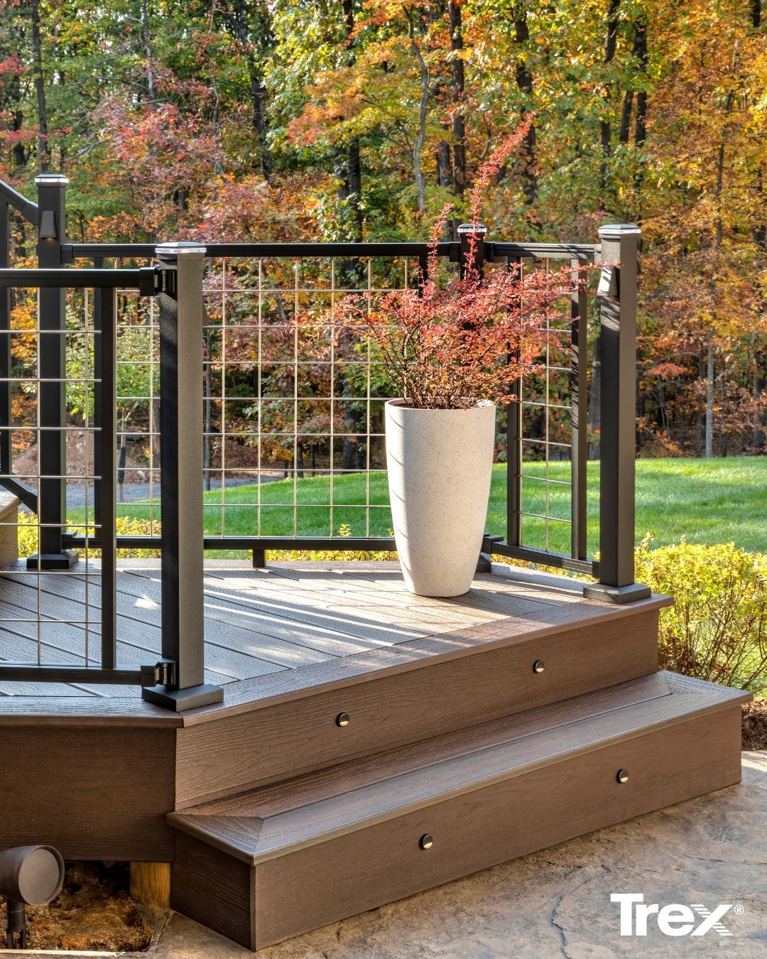 Brown composite deck steps with a black metal railing and a large white planter, set against a blurred autumn background.