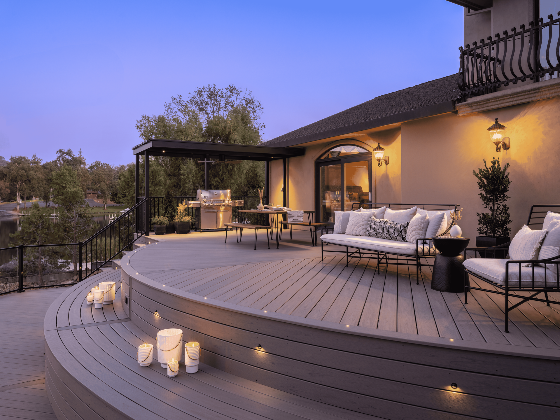 A curved, gray-toned outdoor deck at dusk, featuring lounge seating, an outdoor grill area, and warm step lighting.
