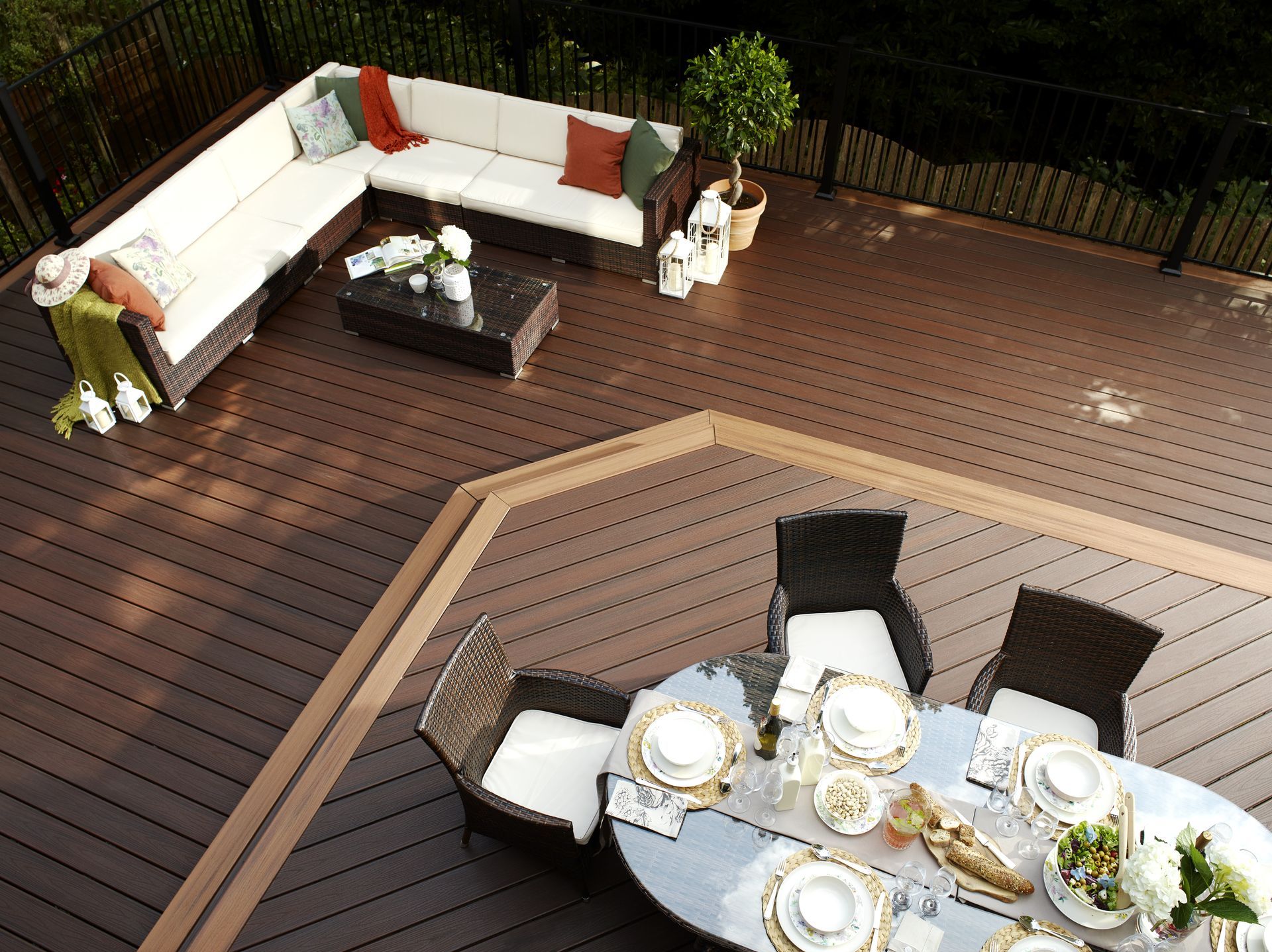 An outdoor deck with a dark rattan sectional sofa and a glass-topped dining set, surrounded by a metal railing.