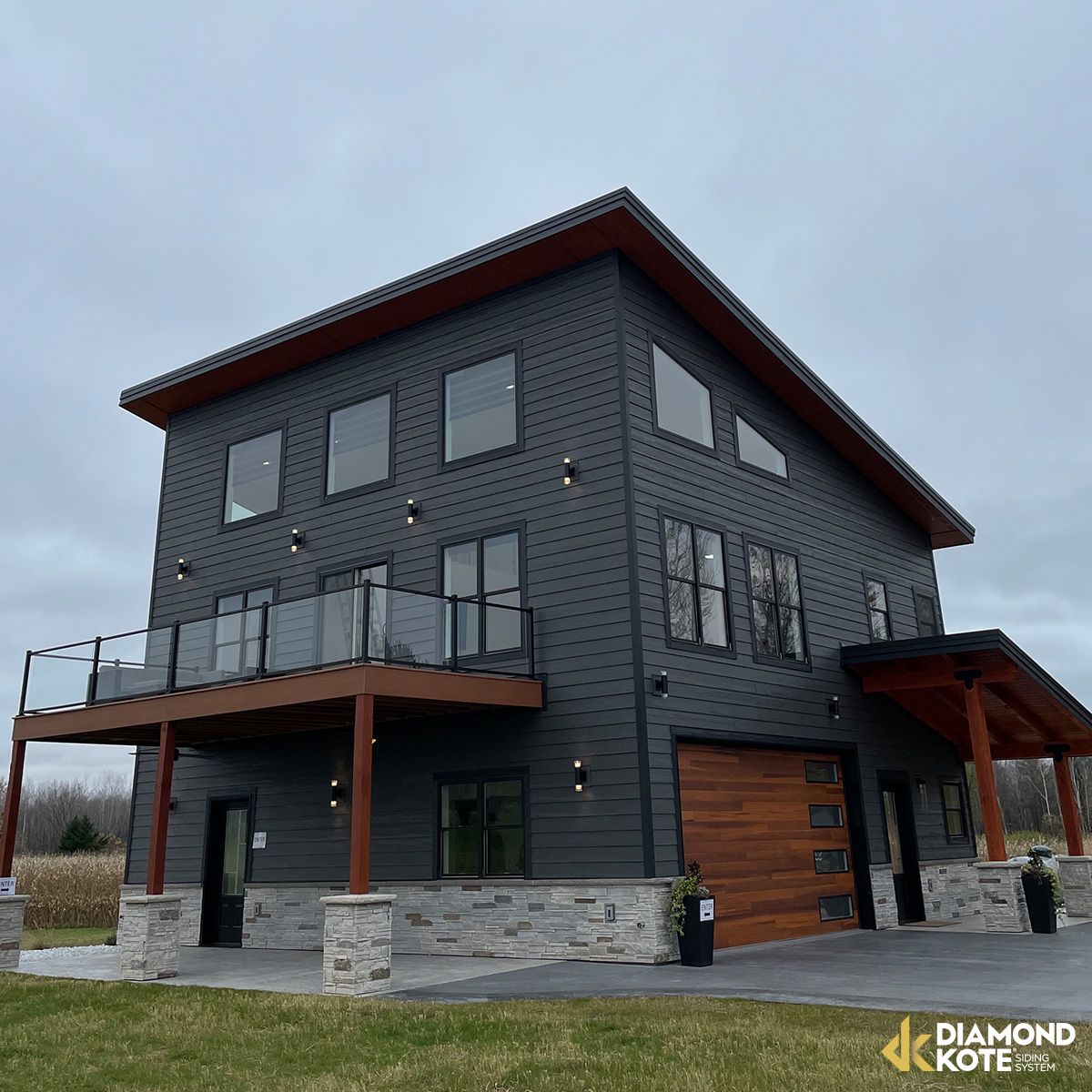 Modern two-story dark-gray house with wood accents, stone base, balconies, and large windows under an overcast sky