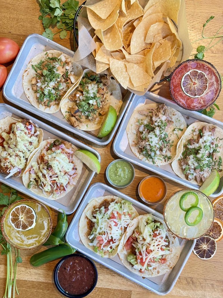 Assorted food trays with rice, sauces, chips, and drinks on a colorful table