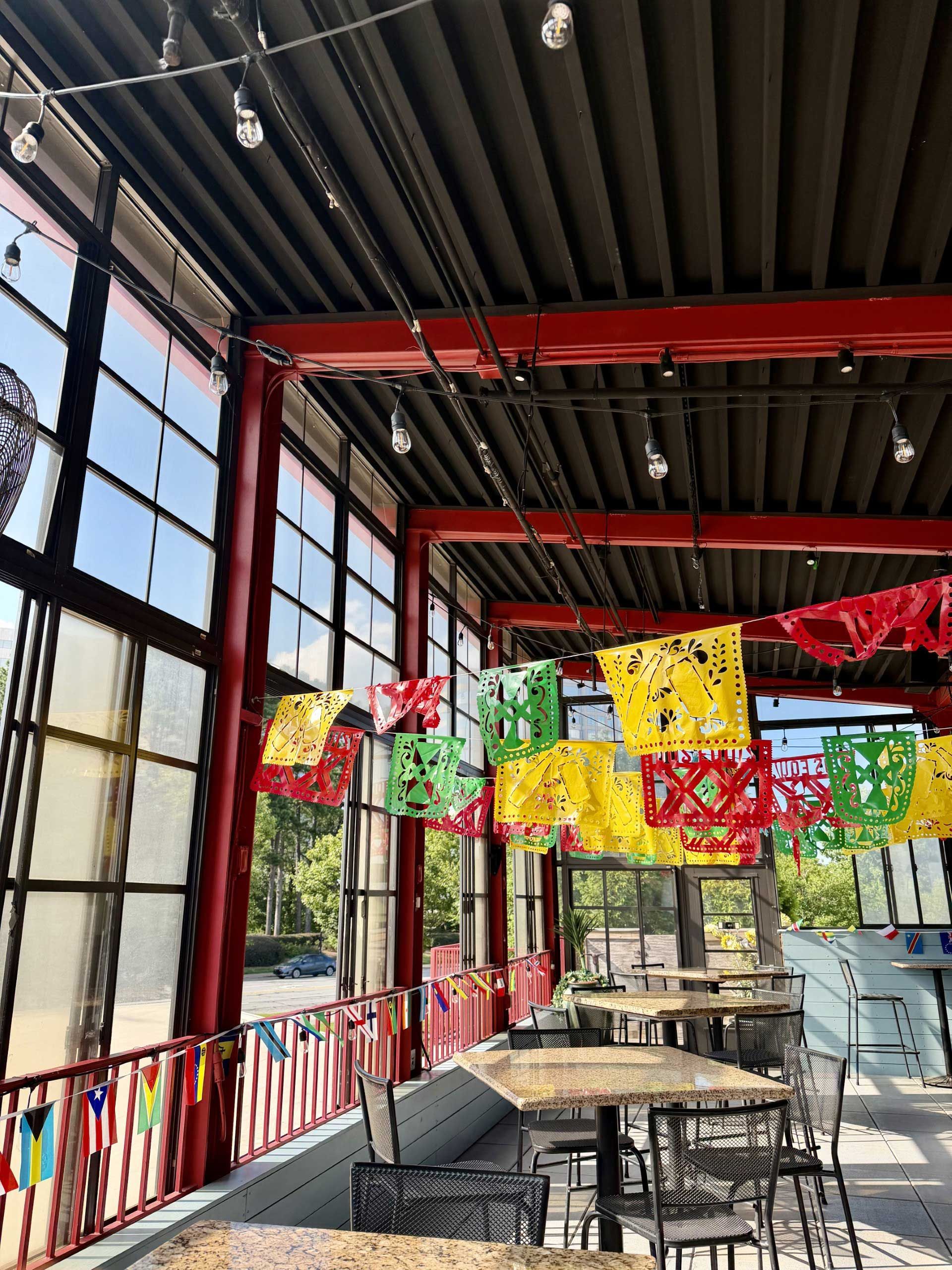 Outdoor rooftop patio with tables, red beams, colorful papel picado, and city views through large windows