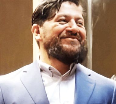 Man with beard smiling, wearing a light blue suit jacket and white shirt.