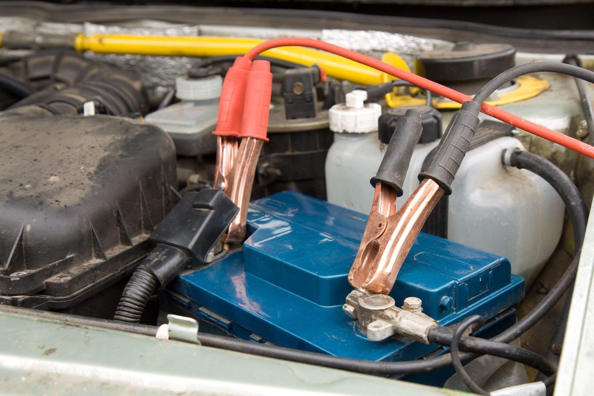 Car battery being jump-started; red and black jumper cables connected to a blue battery in an engine bay.
