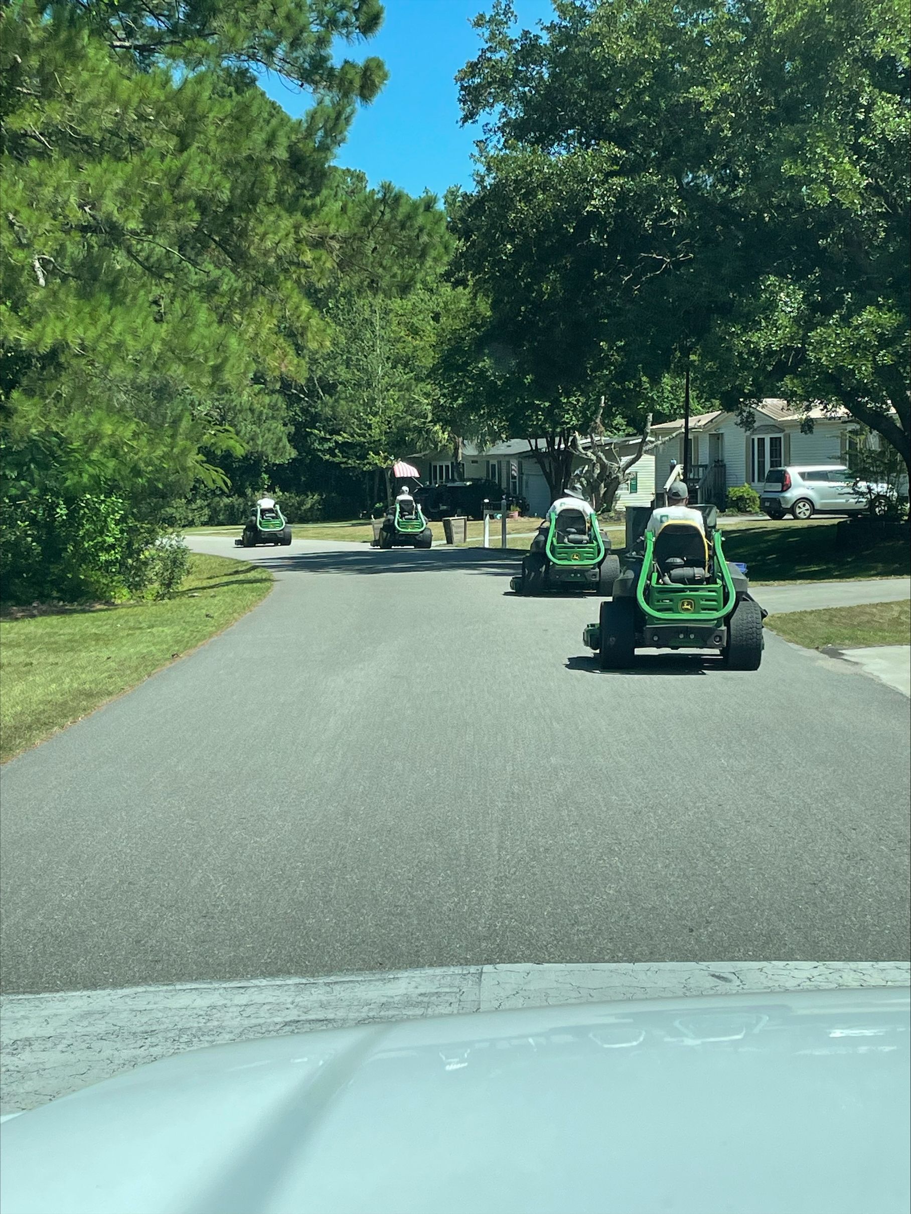Several green lawn mowers on a paved road in a residential neighborhood on a sunny day.
