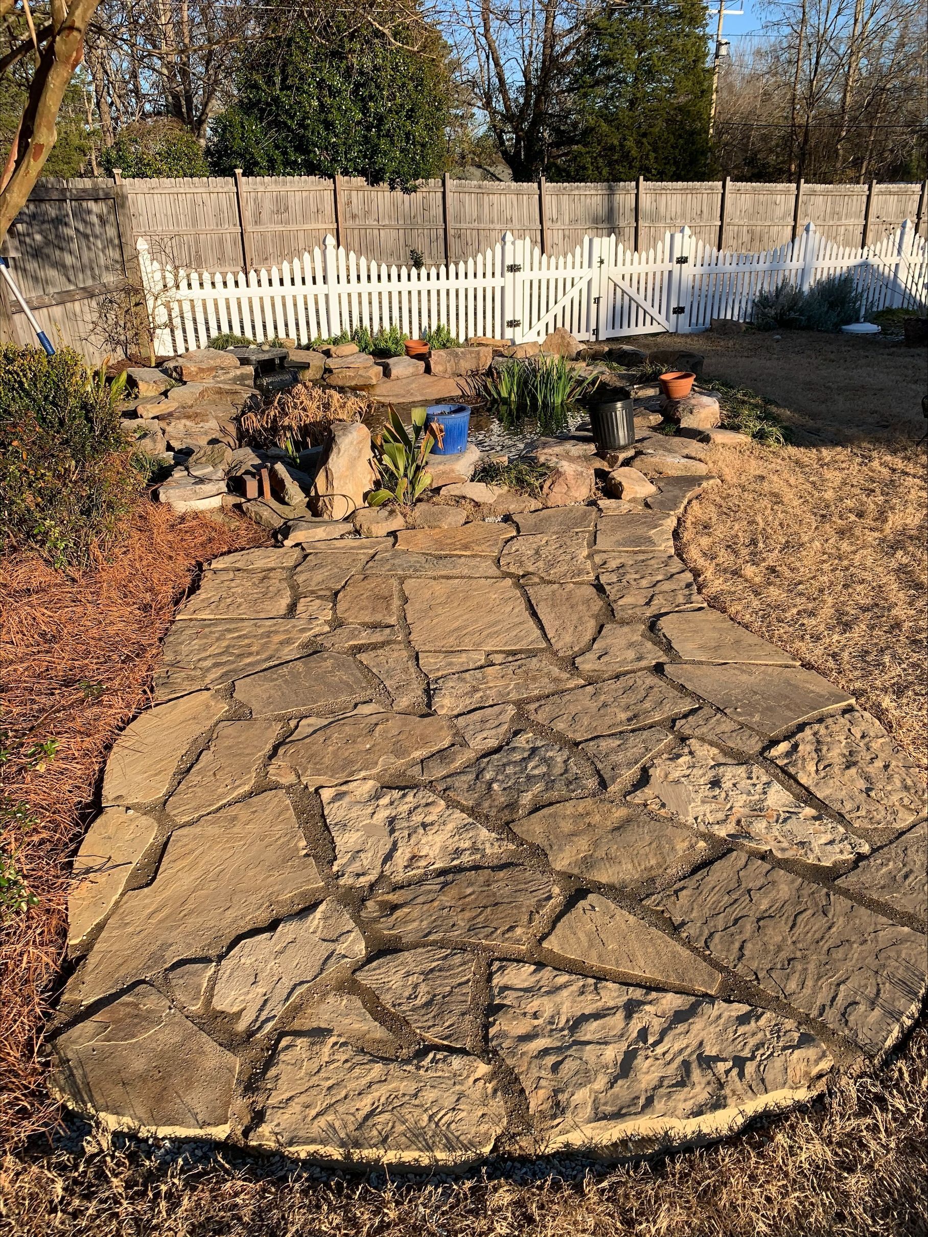 Stone path leads to a garden with a small pond and white picket fence in a backyard.