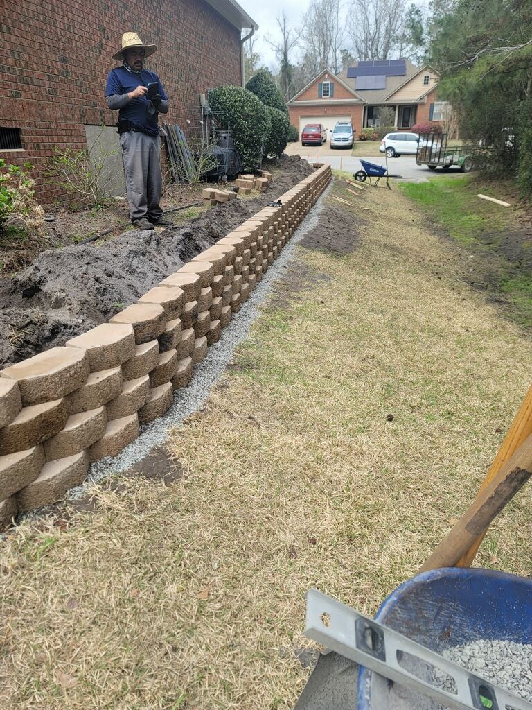Man building a retaining wall with brown blocks, next to yard and house.