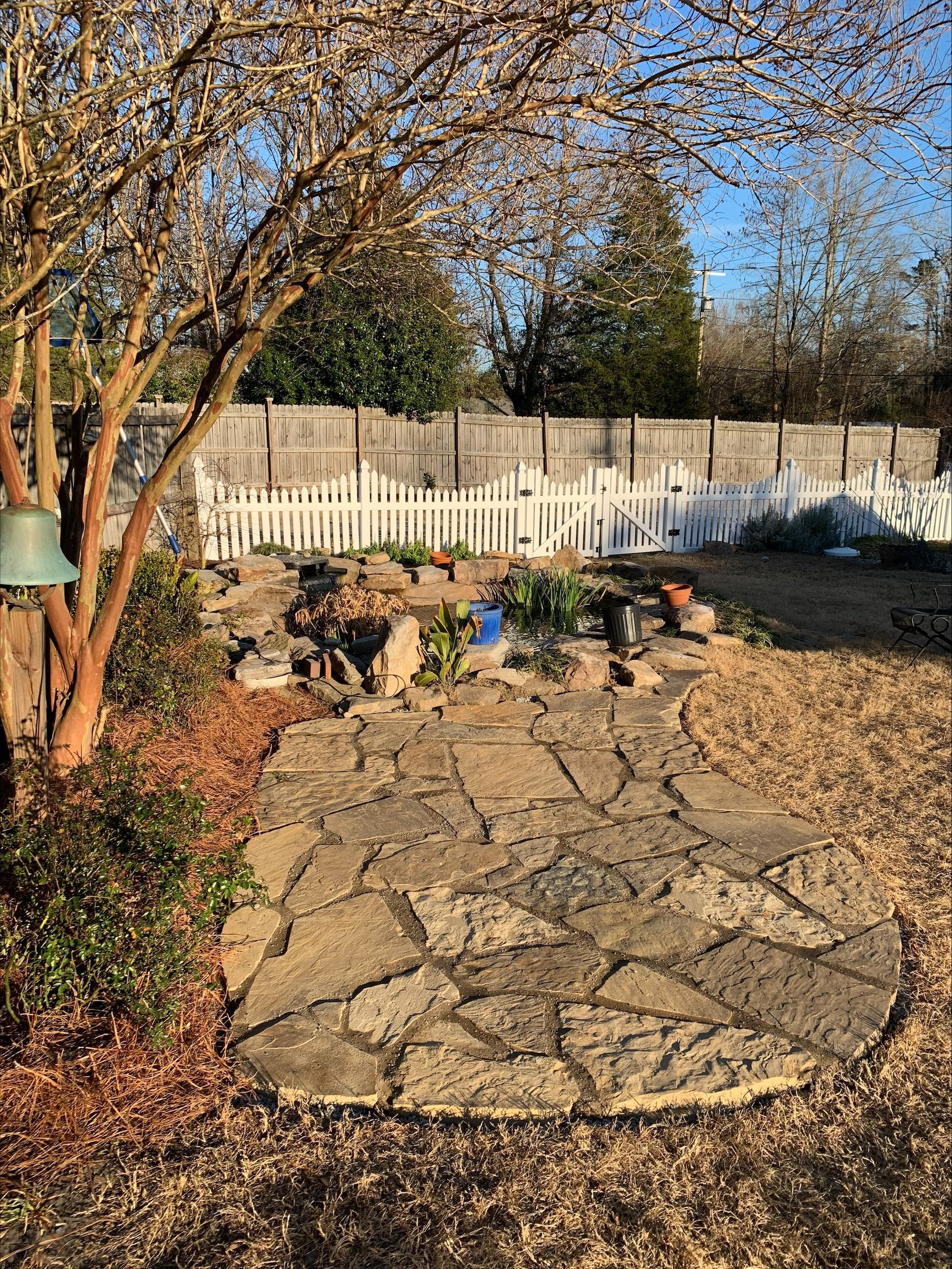 Stone path leads to a garden bed with a small pond; white picket fence, tree.