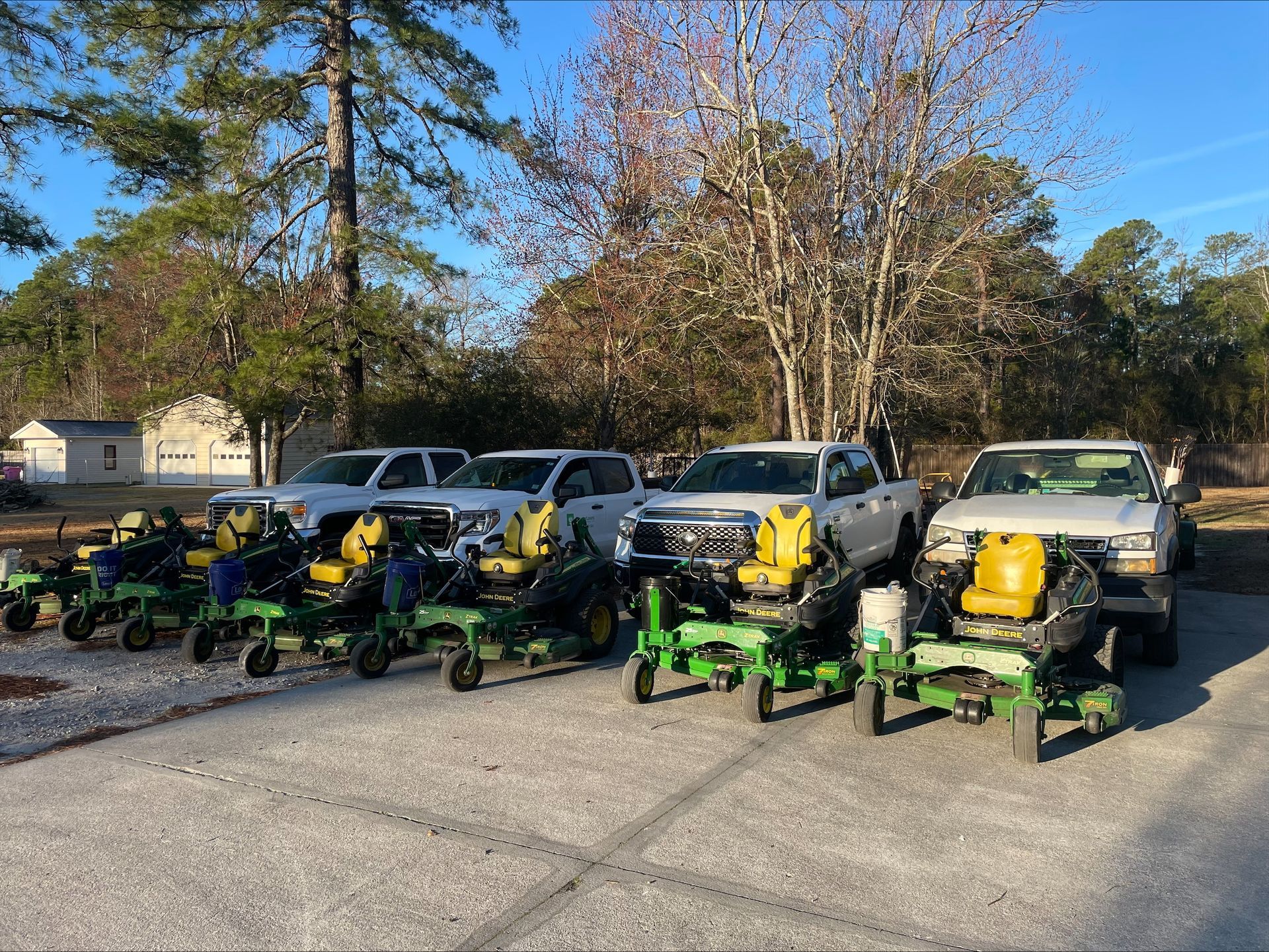 Six John Deere planters lined up in front of white pickup trucks on a gravel lot under a sunny sky.
