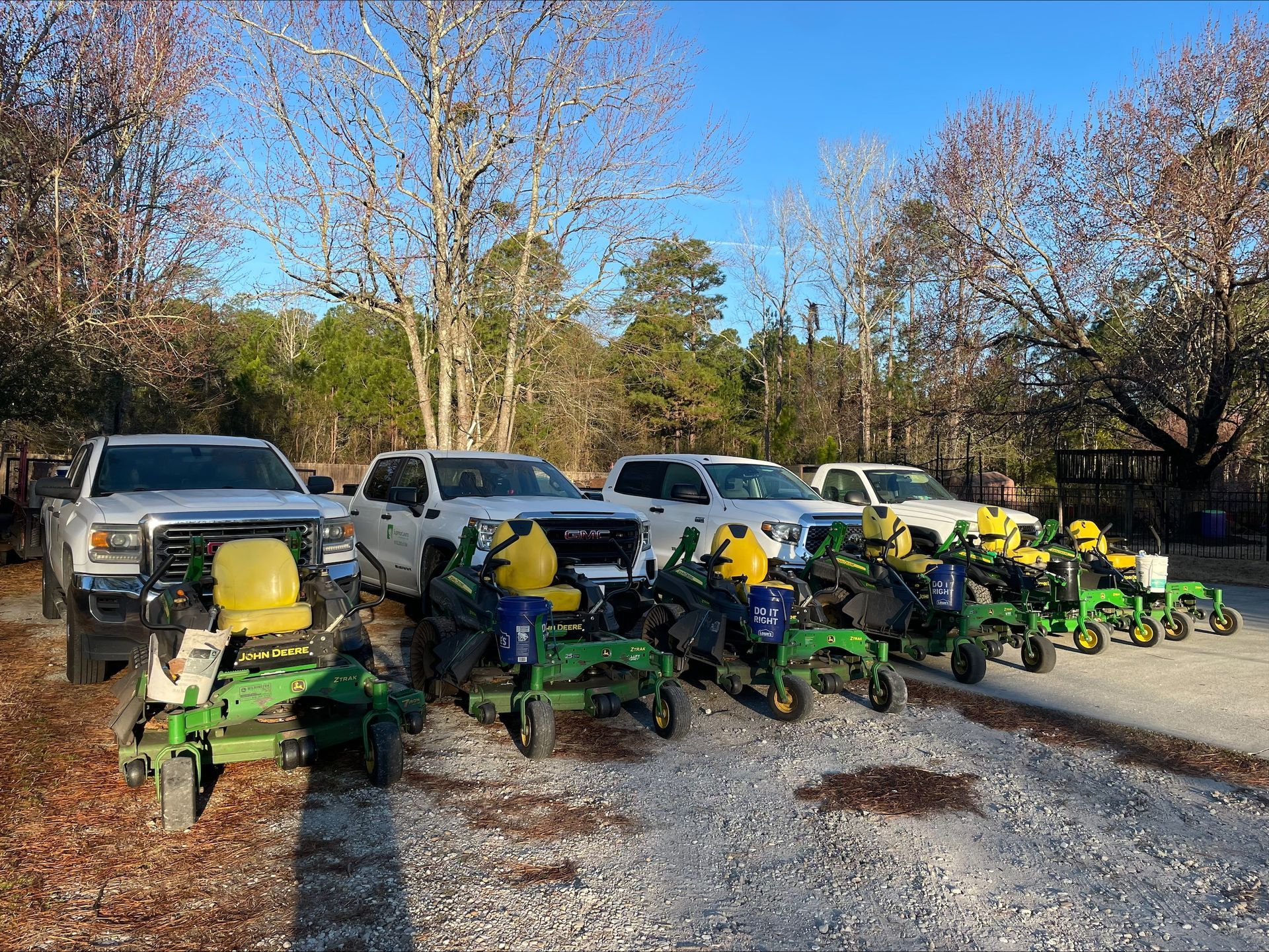 Several white trucks and many green John Deere riding mowers parked outside on a sunny day.