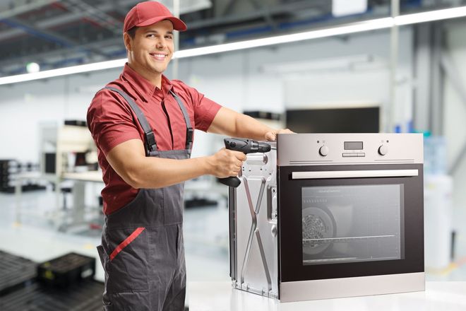 Worker in a red cap smiling while loading a stainless steel oven in an industrial kitchen.