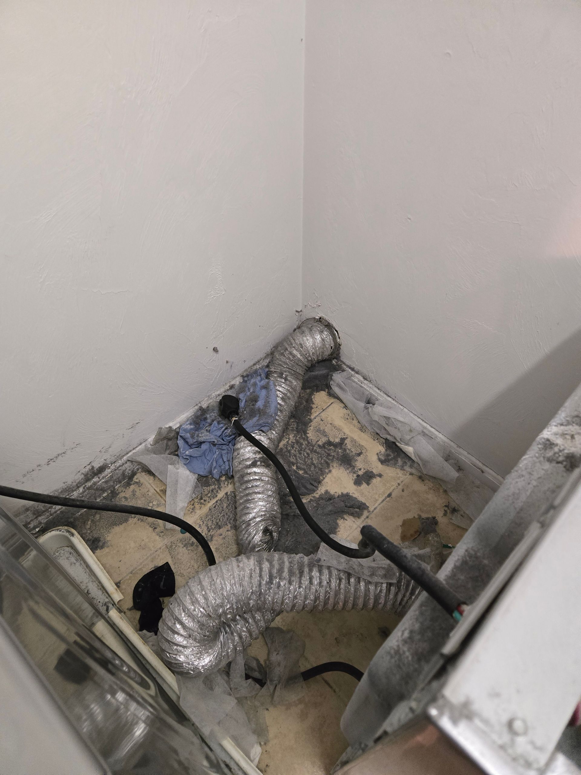 Exposed silver ducting and black cables in a dusty corner behind an appliance.