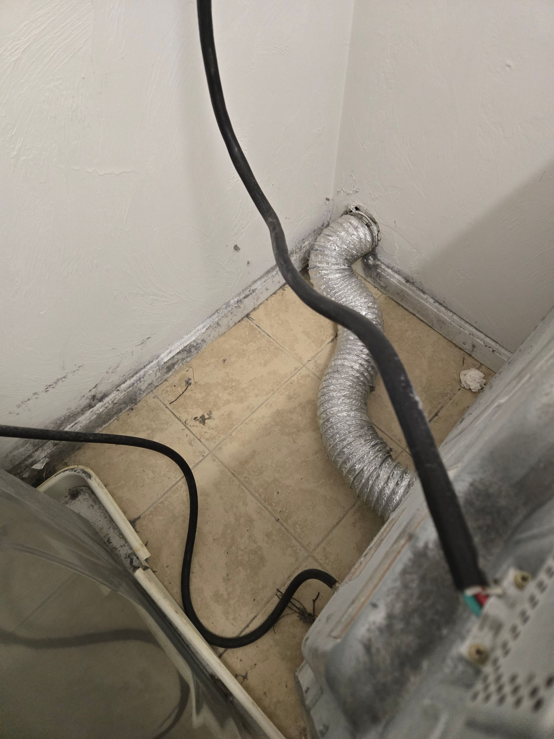 Corner of a room with black and silver cables and a vent hose on a dusty floor.