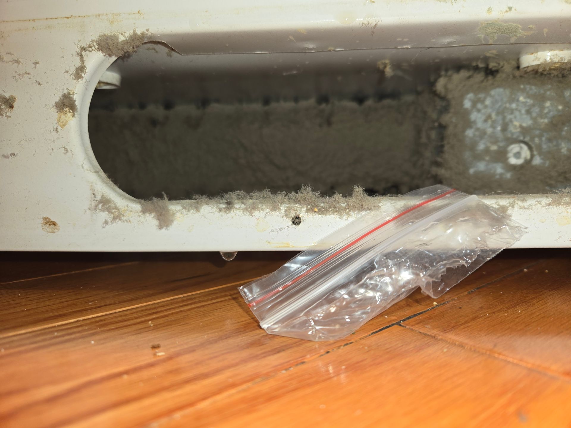 White metal opening above a wooden floor, with a small plastic bag of screws underneath